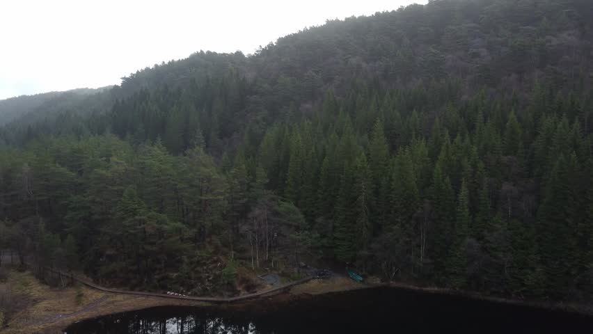 High-angle view flying over a vast coniferous forest on a foggy day, slowly tilting down to reveal a serene mountain lake, a wooden boardwalk, and a canoe resting on the shore