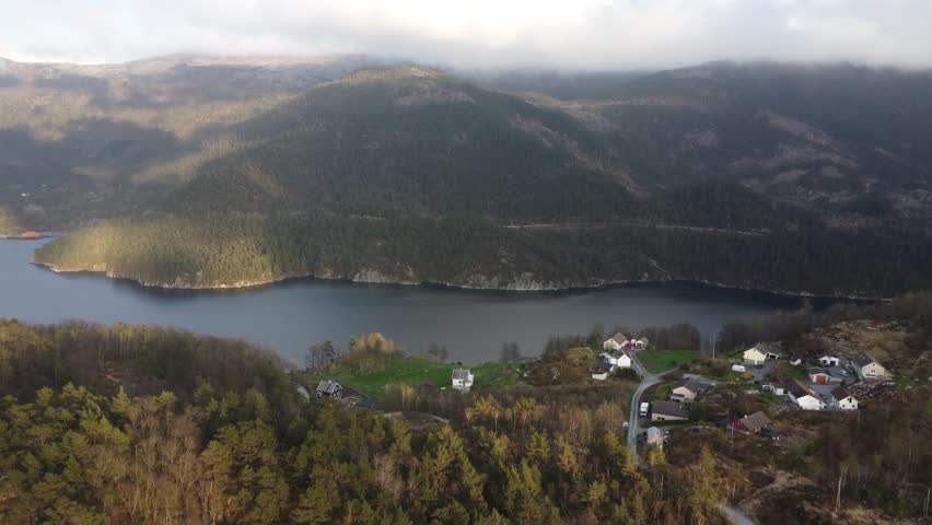 Stunning aerial footage descending towards a picturesque Norwegian village situated on the tranquil shore of a large lake, surrounded by majestic mountains and verdant forests