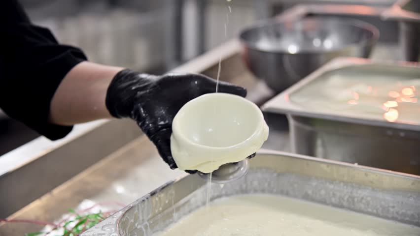 Hands in black gloves shape burrata cheese in a kitchen, with bowls of ingredients and green ropes visible on the countertop