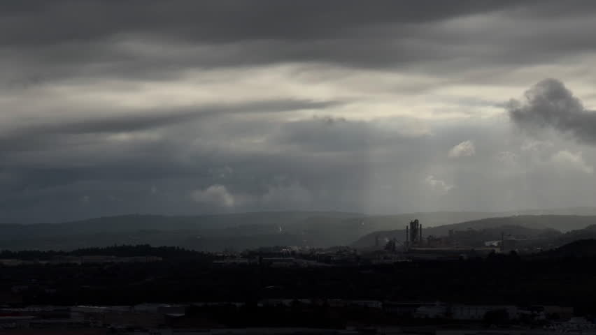 Panoramic view of a cement plant under oppressive gray clouds, with occasional sun breaking through.