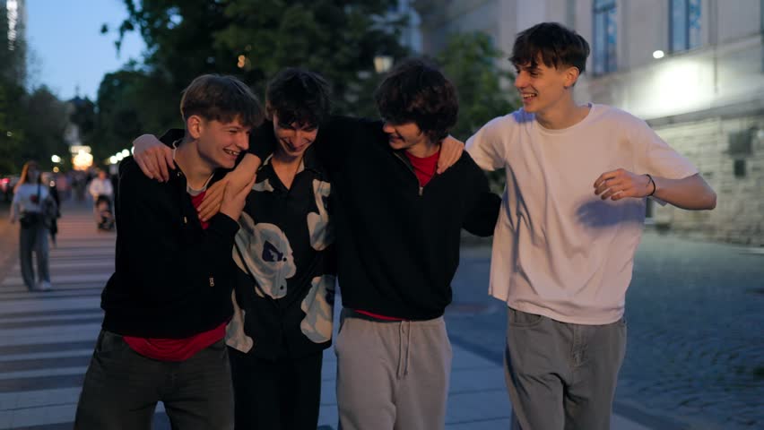 A group of four cheerful caucasian teens in casual outfits walk arm in arm during evening. They enjoy a fun night out in the urban city with a friendly atmosphere.