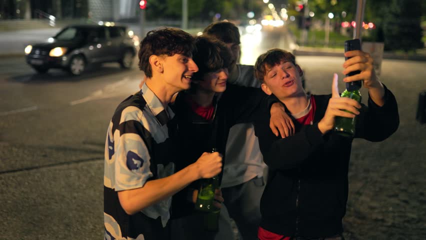 Cheerful young caucasian friends gather close at night to take a self portrait phone photo. The happy companions smile, laugh, and hold green beer bottles at dusk.