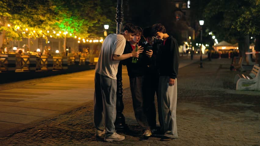 A group of young caucasian friends leans against a street lamp column at night. The companions look closely at a smartphone screen while holding beer bottles.
