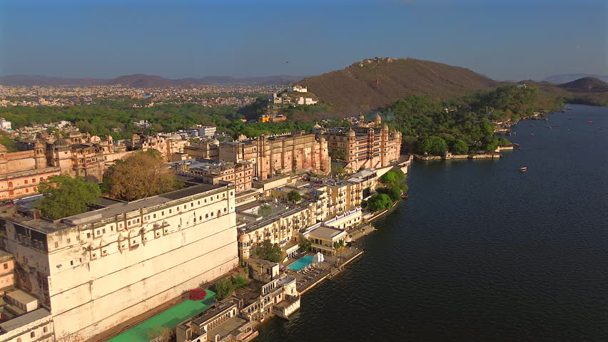 A high-angle wide static shot of the majestic City Palace and luxury heritage hotels along the banks of Lake Pichola, with the urban skyline and Aravalli hills in Udaipur, Rajasthan, India.