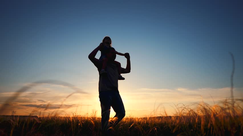 A man carries a girl on his shoulders against a sunset backdrop. The man and girl are silhouetted by the golden light as they stand in a grassy field.