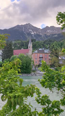 Beautiful scenic view of a church tower, colorful buildings and alpine mountains above the Inn River in Innsbruck Austria. Peaceful European landscape in Tyrol surrounded by green trees, dramatic clouds and snowy mountain peaks. Atmospheric Austrian travel destination combining architecture and nature in the Alps.