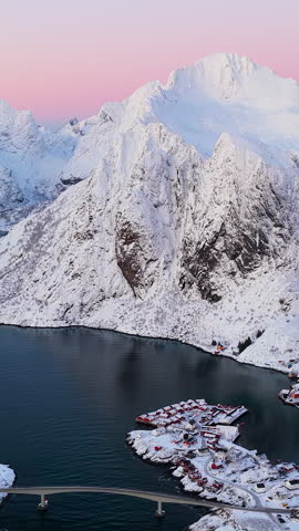 Cinematic aerial drone shot of snowcovered peaks and traditional red cabins in Lofoten, Norway during pink golden hour.
