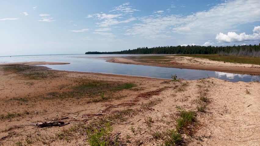 Aerial view of an empty sandy shoreline along a calm lake, with gentle waves, shallow water and distant tree-lined horizon under a partly cloudy sky