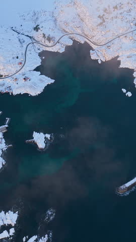 Cinematic vertical aerial shot of the rugged Lofoten islands coastline in Norway covered in snow with deep blue arctic water.