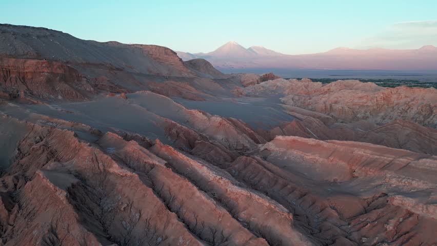 Forward flight over Valle de la Luna vast eroded rock formations at dusk with snow-capped Licancabur volcano glowing pink in twilight sky. San Pedro de Atacama, Chile