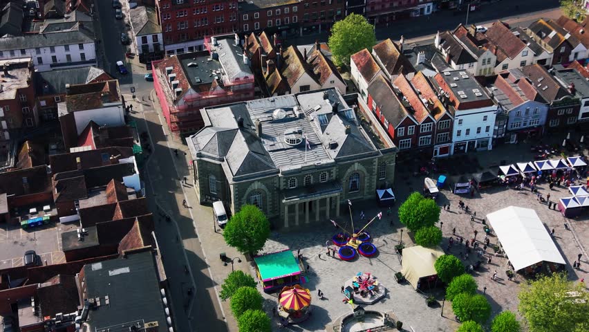 Aerial drone footage looking down over Salisbury Guildhall and Market Square during a St George’s Day event, with fairground rides, market stalls, crowds, historic buildings, streets, and rooftops