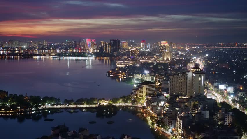 Night aerial cityscape of West Lake and Thanh Nien Road with illuminated urban skyline, reflective waterfront, and vibrant Hanoi evening atmosphere.