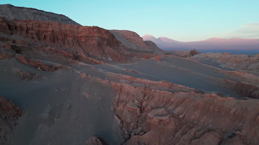 Low altitude flight through Valle de la Luna narrow canyon between eroded rock formations at dusk with snow-capped Licancabur volcano in pink twilight sky. San Pedro de Atacama, Chile