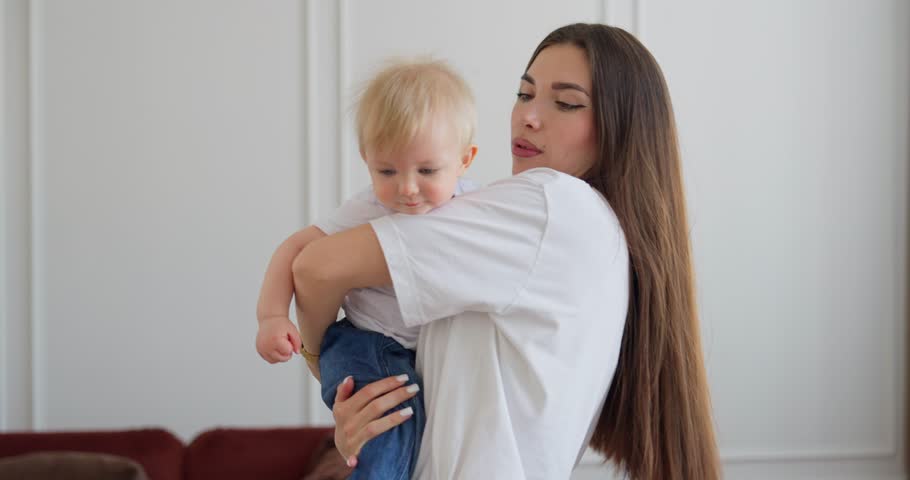 Young mother tenderly holding her blond baby in a bright home interior, sharing a quiet affectionate parenting moment with gentle care