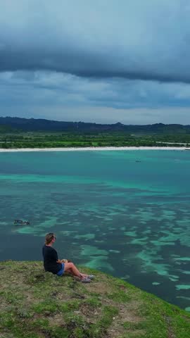 Woman on cliff viewpoint overlooking ocean, dark top and shorts, grassy bluff with panoramic turquoise water and visible reef patterns, distant sandy shoreline under heavy overcast clouds, Lombok