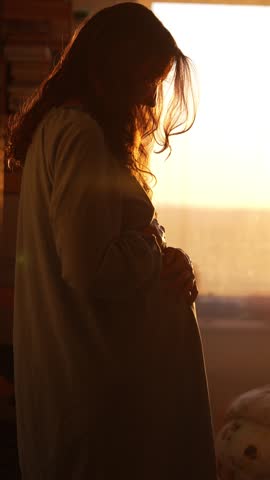Backlit pregnant woman affectionately caressing her belly, standing by a window and looking out at the morning sun, feeling the first movements of her child in a moment of connection