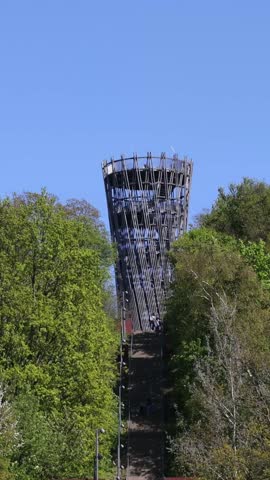 People climbing a steep staircase to an architectural observation tower on a hill surrounded by trees