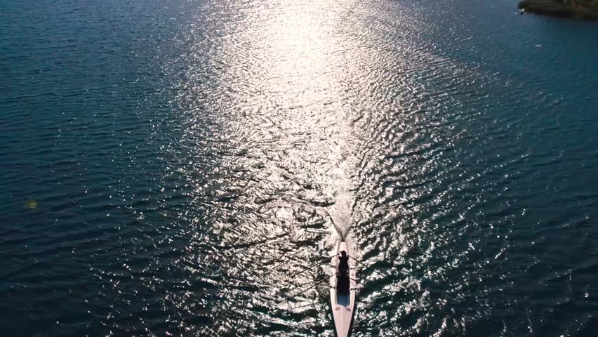 Drone shot of rowers moving across sunlit lake water with bright reflections and ripples. Peaceful aerial scene showing outdoor sport, endurance, teamwork, freedom and nature.