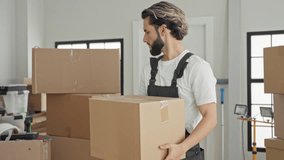 Man in apron carrying cardboard box in building with stacked boxes, drill and worklight visible, looking toward camera while handling package; moving day determination. - Powered by Shutterstock - Get 15% off with code: PIKWIZARD15