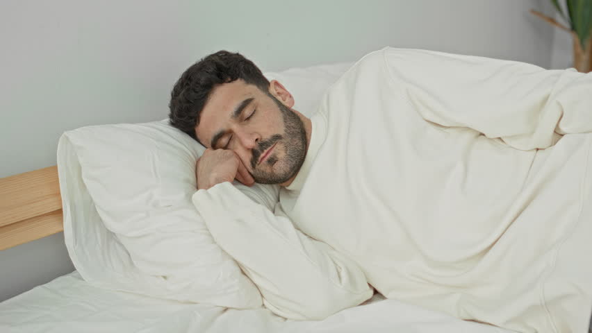 Man with hand on stomach reclining on bed in a building, eyes closed and facial grimace showing pain; discomfort fatigue.