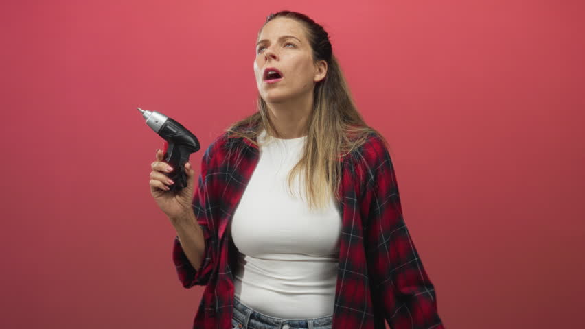 Woman holding cordless screwdriver covers one eye with her hand and looks weary while wearing plaid shirt and white tee in studio; irritation caution.