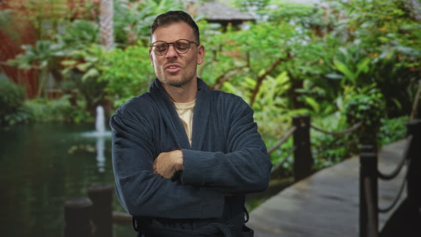 Man in robe wearing glasses with arms crossed on a forest boardwalk by a pond and fountain, looking resolute; quiet determination.