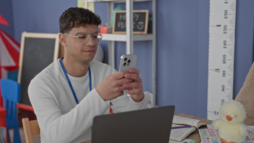 Young man teacher holds smartphone with both hands and smiles while seated at laptop in preschool classroom; cheerful engagement.