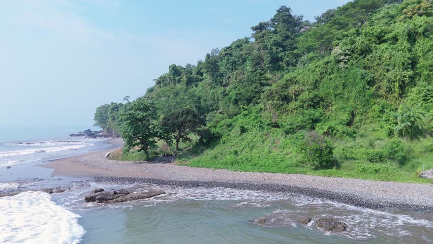 Aerial view of ocean waves crashing onto rocky beach coastline in Indonesia with tropical scenery