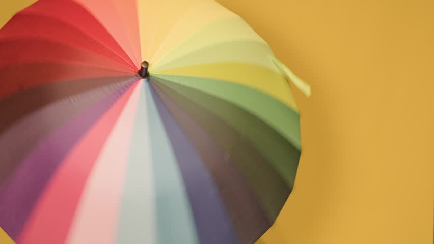 Cheerful young woman with curly hair holding a large, colorful rainbow umbrella. She is smiling and posing against a bright yellow background
