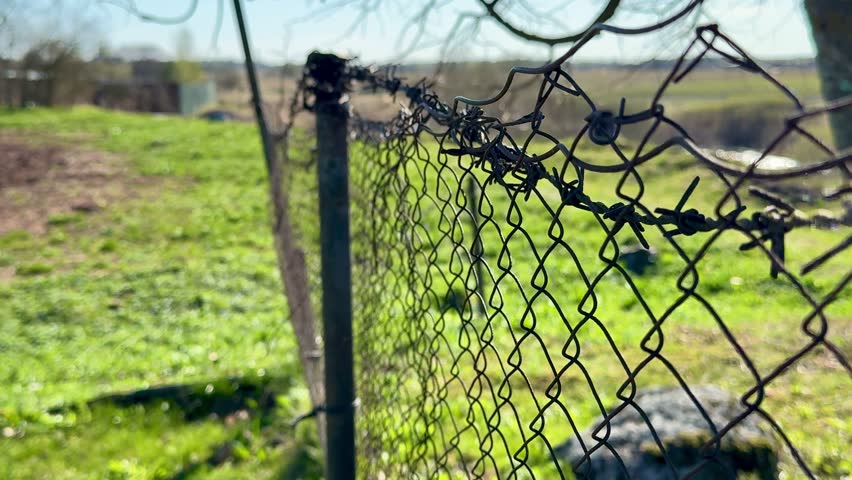 Rusty chain fence on garden plot under bare tree branches, leaning metal post and old pipe casting long shadow across fresh spring.
