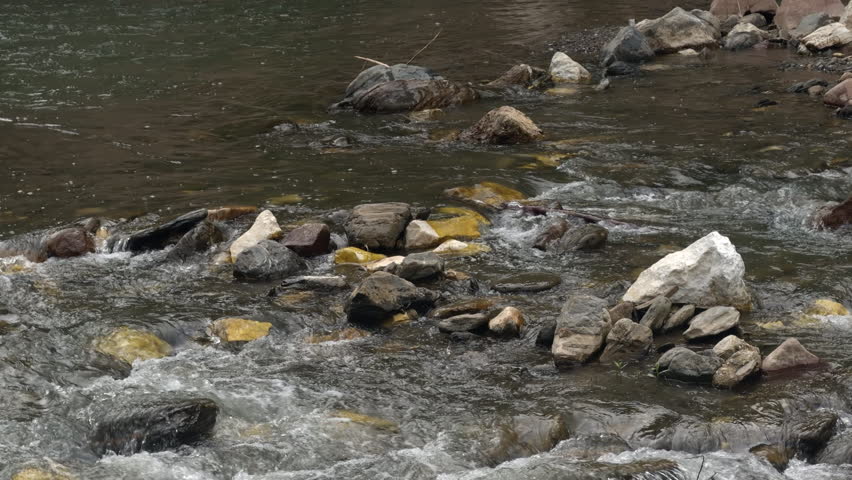 Clear river water flowing around natural rocks and stones in a peaceful outdoor setting. Scenic freshwater stream with gentle current and natural landscape texture.