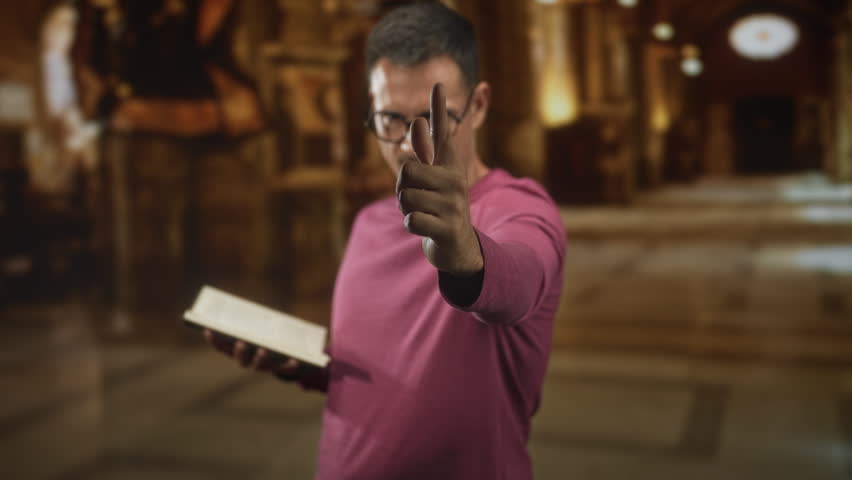 Young hispanic man with moustache and glasses holds an open book and points finger at camera inside ornate building; confidence focus.