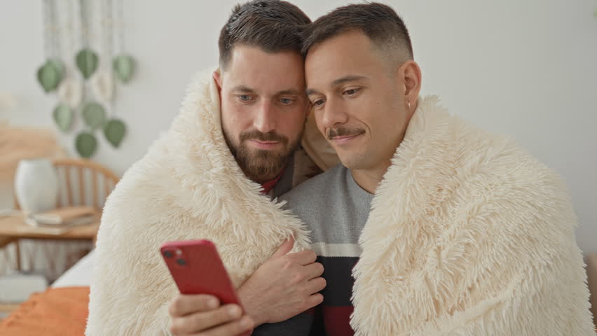 Two men cuddling under a cream blanket, one holding a red smartphone and kissing the other