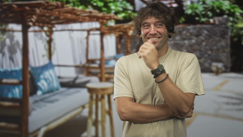 Hispanic man with hand covering mouth and crossed arms in building cabana lounge wearing a watch and bracelets near daybed and pillows; amused vacation moment.