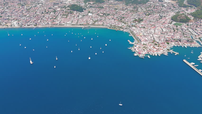 Marmaris, Turkey. A sweeping aerial panorama captures the broad natural harbor that has drawn sailors since antiquity, its deep blue waters dotted with dozens of white yachts and sailboats at anchor. The densely built resort town spreads along the curving shoreline, with terracotta rooftops and low-rise blocks giving way to sandy beaches and a busy marina. Pine-clad mountains rise behind the urban strip, their green slopes contrasting sharply with the turquoise bay. The scene is bathed in bright midday summer light under a clear sky, conveying the atmosphere of a thriving Mediterranean destination. The bay