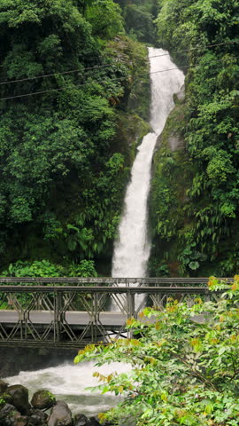 Scenic view of a majestic waterfall flowing through a dense green rainforest, with water rushing under a steel bridge into a turbulent river below, creating a stunning natural landscape