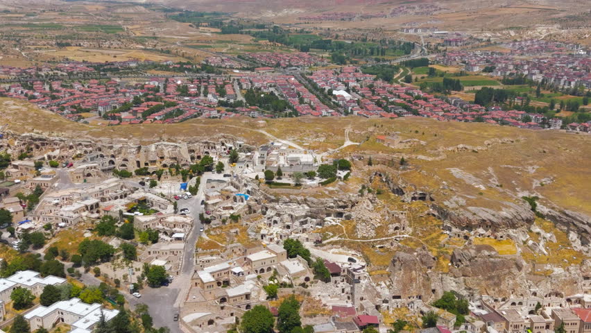 Urgup, Nevsehir, Turkey. Ancient rock-carved settlement and cave dwellings carved into soft volcanic tuff cliffs, an aerial drone view that captures the dramatic contrast between the historic troglodyte architecture and the modern urban expansion of the new town beyond. The landscape reveals the distinctive honeycomb of human-made caves, arches, and terraces that have been inhabited since the Bronze Age, hewn from the region
