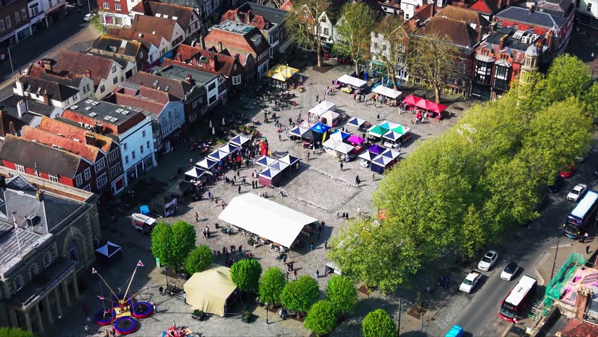 Aerial drone footage looking down over Salisbury Market Square during a St George’s Day event, showing market stalls, crowds, fairground rides, trees, traffic, the Guildhall, historic buildings,