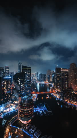 Vertical wide angle timelapse of Dubai Marina skyline at night with moving clouds, traffic crossing bridge and boats cruising through illuminated waterfront canal in UAE
