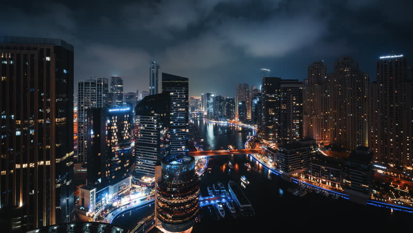 Wide angle timelapse of Dubai Marina skyline at night with moving clouds, traffic crossing bridge and boats cruising through illuminated waterfront canal in United Arab Emirates