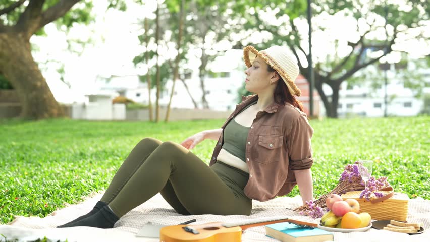 Beautiful woman sitting on a picnic blanket in a sunny park, feeling the summer heat and using her straw hat as a fan to cool herself while enjoying a relaxing day outdoors