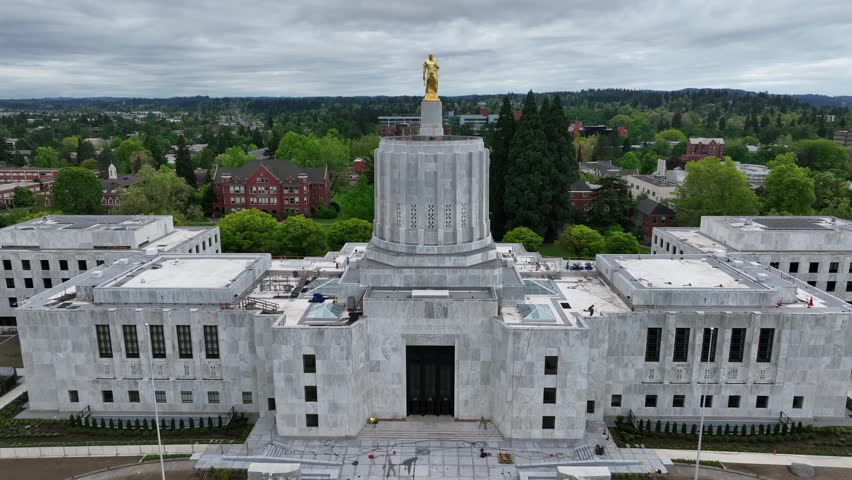 Oregon State Capitol building in Salem with white marble facade and Art Deco dome with golden statue surrounded by lush green trees under overcast sky during spring in United States. Aerial rise wide