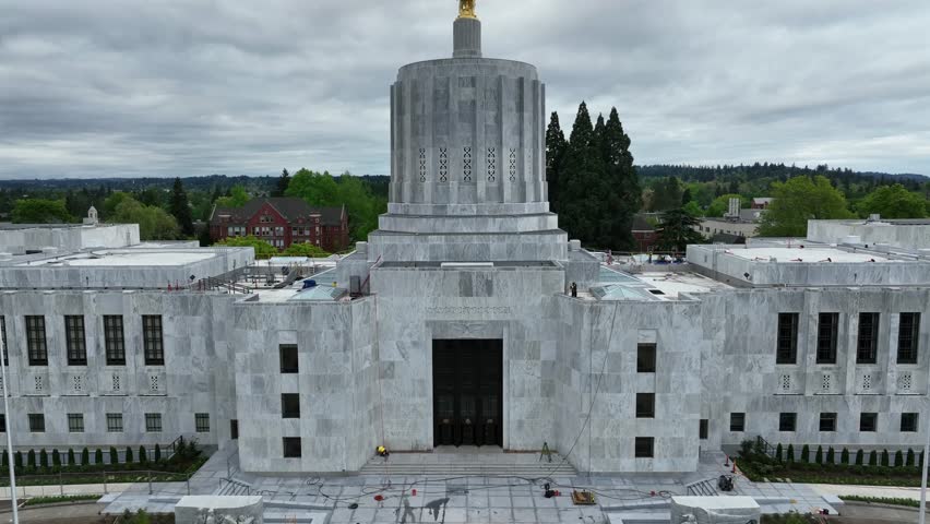 Oregon State Capitol in Salem with gold Pioneer statue atop Art Deco dome with worker on roof and green trees under overcast sky during spring daytime in American Pacific Northwest. Aerial rising view