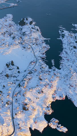 High angle aerial shot of a traditional Norwegian coastal village in Lofoten covered in snow during winter sunset, featuring a small harbor and winding roads.