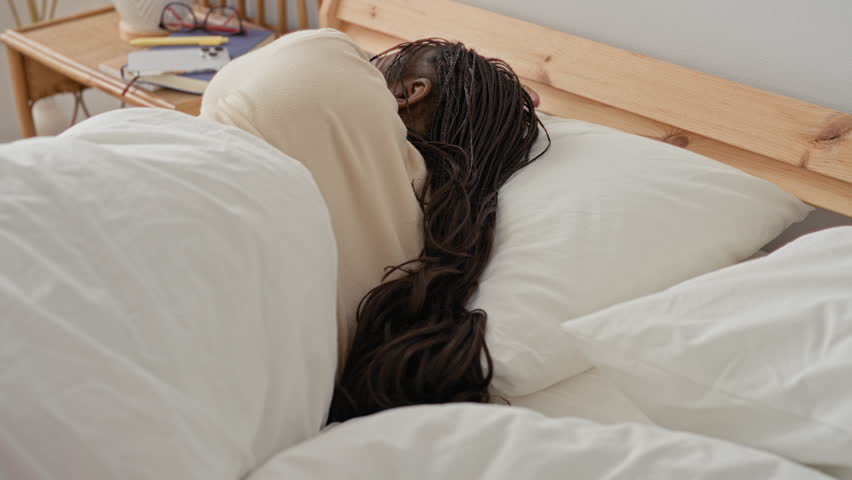 Woman touching her shoulder with a hand while turning on a bed near a window, vase, books and pampas grass in a bedroom building; relaxation.