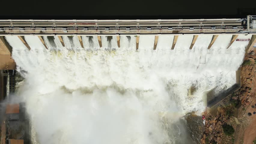 Overhead view: white water rushes through open sluice gates of a concrete dam wall