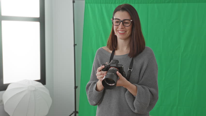 Woman holding dslr camera, hand adjusting glasses and smiling while framing shot in studio; confidence creativity.