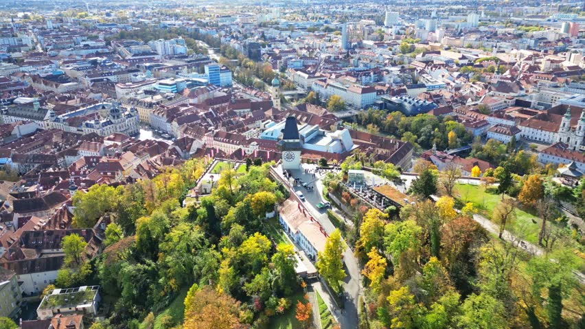 Aerial view of Schlossberg clock tower surrounded by historic red rooftops and autumn foliage in Graz, Steiermark, Austria.