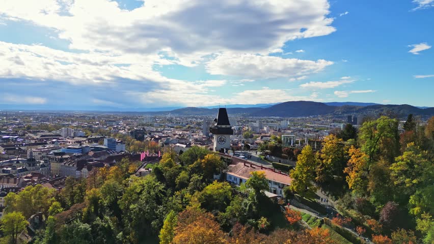 Aerial view of the historic Schlossberg hill and clock tower surrounded by autumn trees and the city skyline in Graz, Styria, Austria.