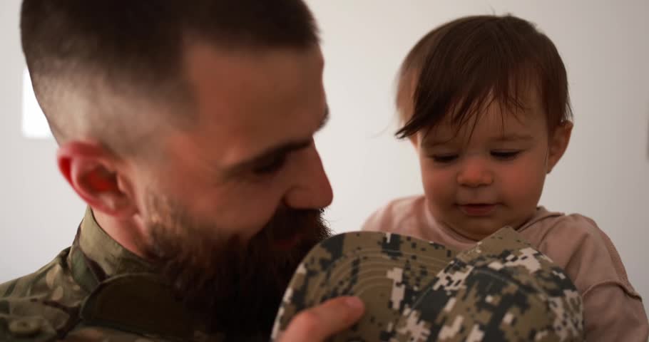 American soldier man in military uniform hugs his little son at home while baby boy playing with his hat - Emotional and happy homecoming concept
